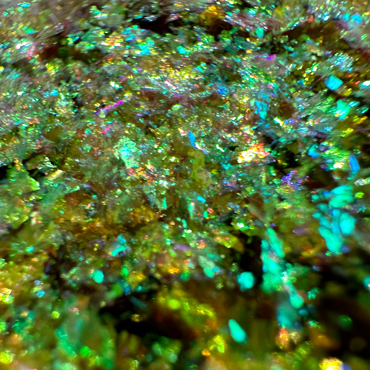 Close-up of iridescent glitter flakes with a holographic finish, showing shifting colors of green, blue, purple, and gold.